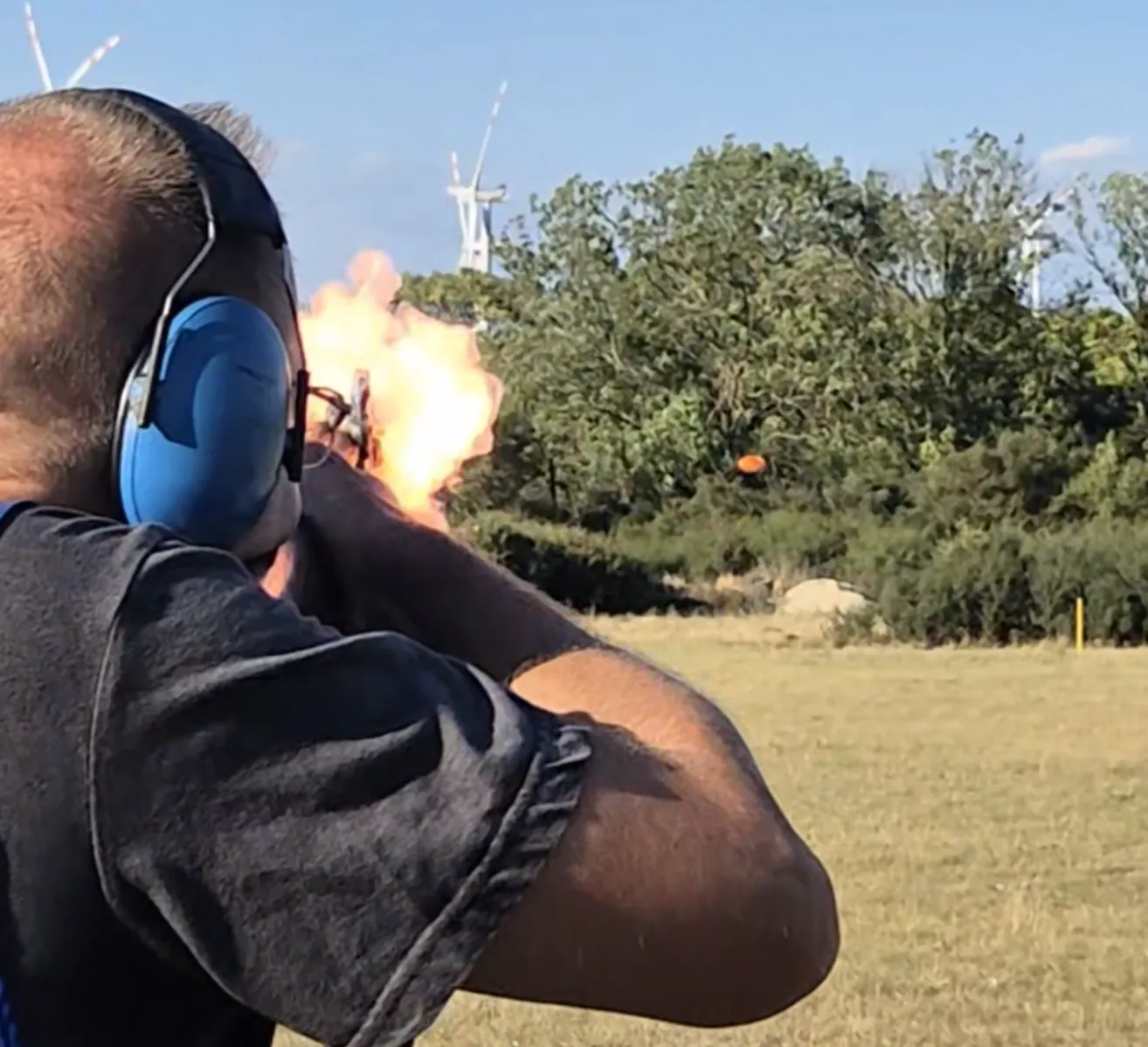 Ein Mann in einem schwarzen Shirt und blauem Gehörschutz schießt eine Waffe im Freien ab, mit einer hellen Explosion im Hintergrund.