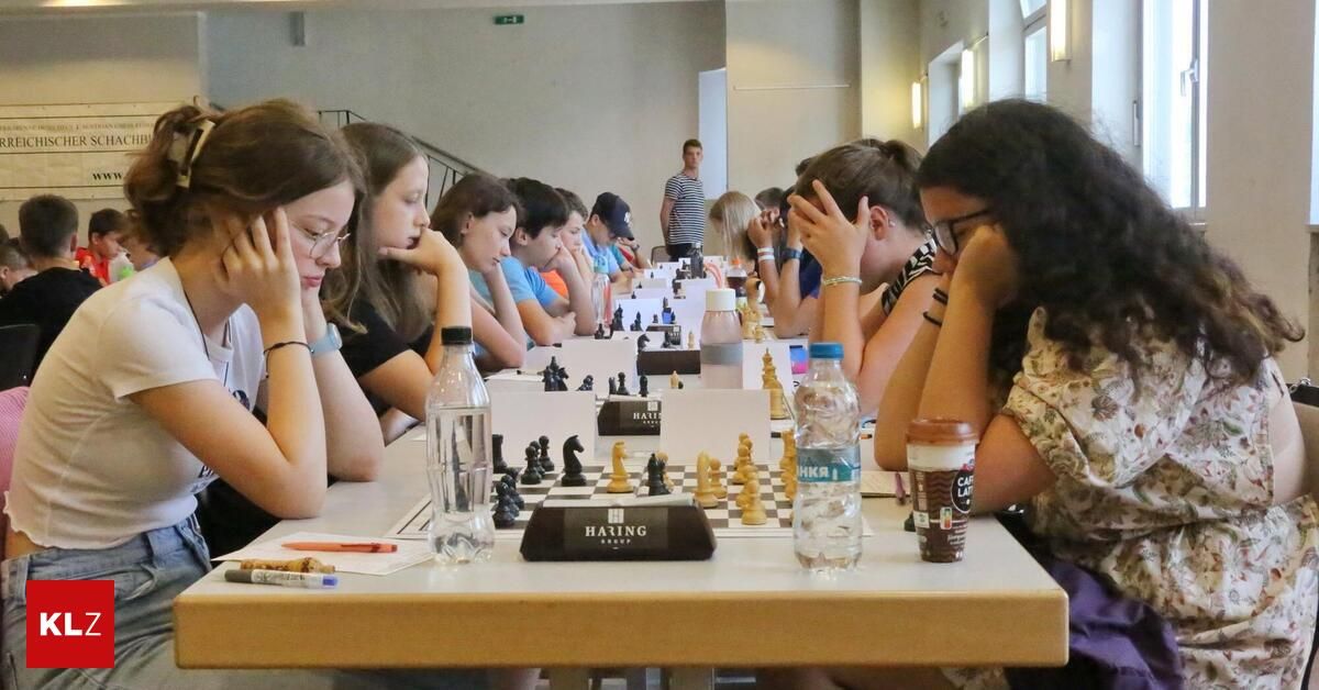 A group of young people are playing chess in a classroom. Each player has a chessboard in front of them, and they are all focused on their games. A man stands behind them, observing.