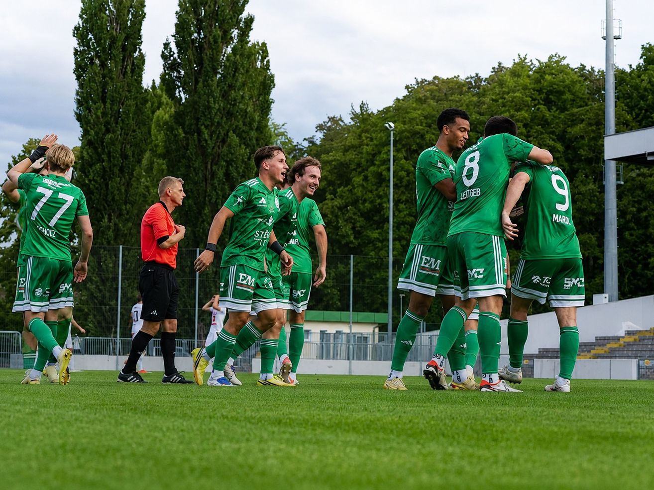 Soccer players in green uniforms celebrate on the field with a referee in red. Trees and a fence are in the background.