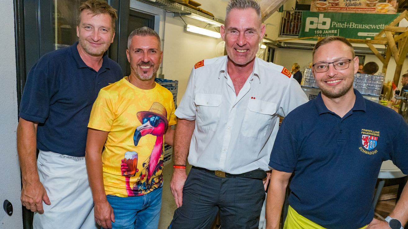 Vier Männer stehen zusammen in einem Raum. Der Mann auf der linken Seite trägt ein gelbes Shirt mit einem Flamingo und einem Hut. Der Mann neben ihm trägt ein weißes Shirt mit roten Abzeichen. Der Mann auf der rechten Seite trägt ein blaues Shirt.