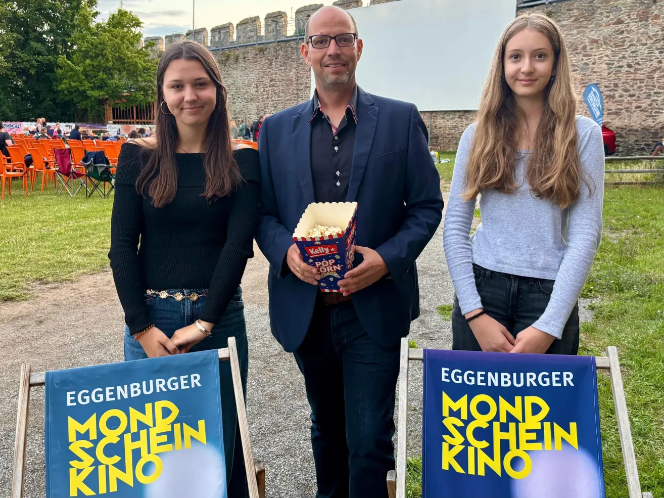 Three individuals stand in front of a movie poster, with the man holding a box of popcorn. The woman on the left wears a black top, while the woman on the right wears a gray top. Behind them is a backdrop of a stone wall and a large screen.