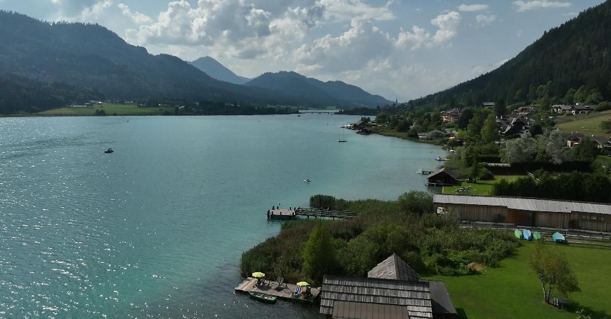 Ein ruhiger See mit einem Steg, Bergen im Hintergrund und einem klaren Himmel mit Wolken.