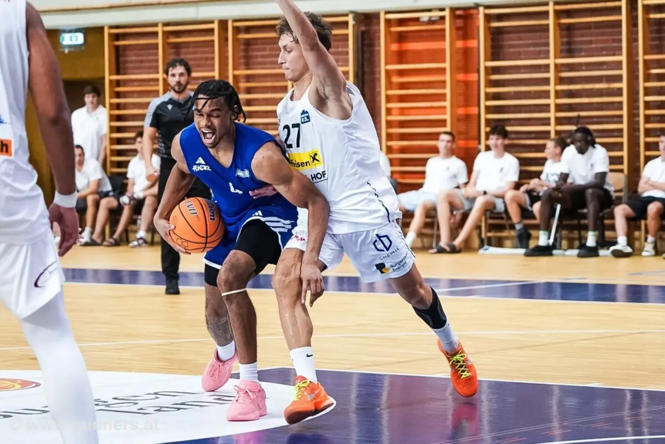 Two basketball players are engaged in a game on an indoor court. One player in a blue jersey is dribbling the ball while the other in a white jersey tries to block him. Behind them, spectators are watching the game.