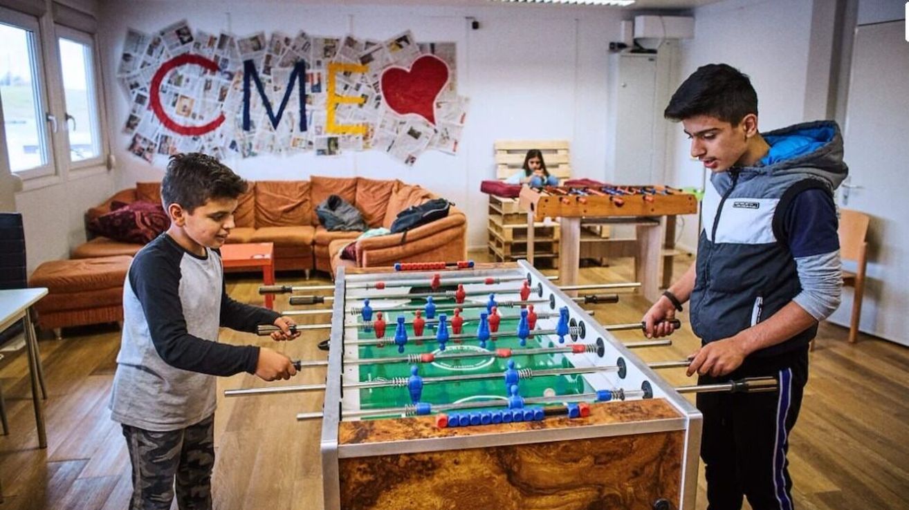 Two boys play table soccer in a room with a couch, a wooden table, and a wall covered in newspaper. A girl sits on a stack of wooden pallets.