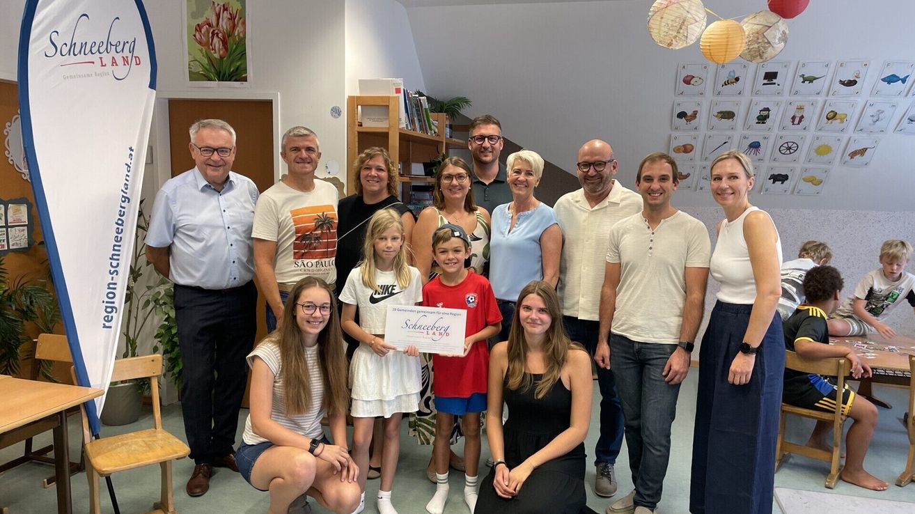 A group of people, including adults and children, are posing for a photo. A girl in the middle holds a certificate. The room has a white wall with a staircase and a bookshelf.