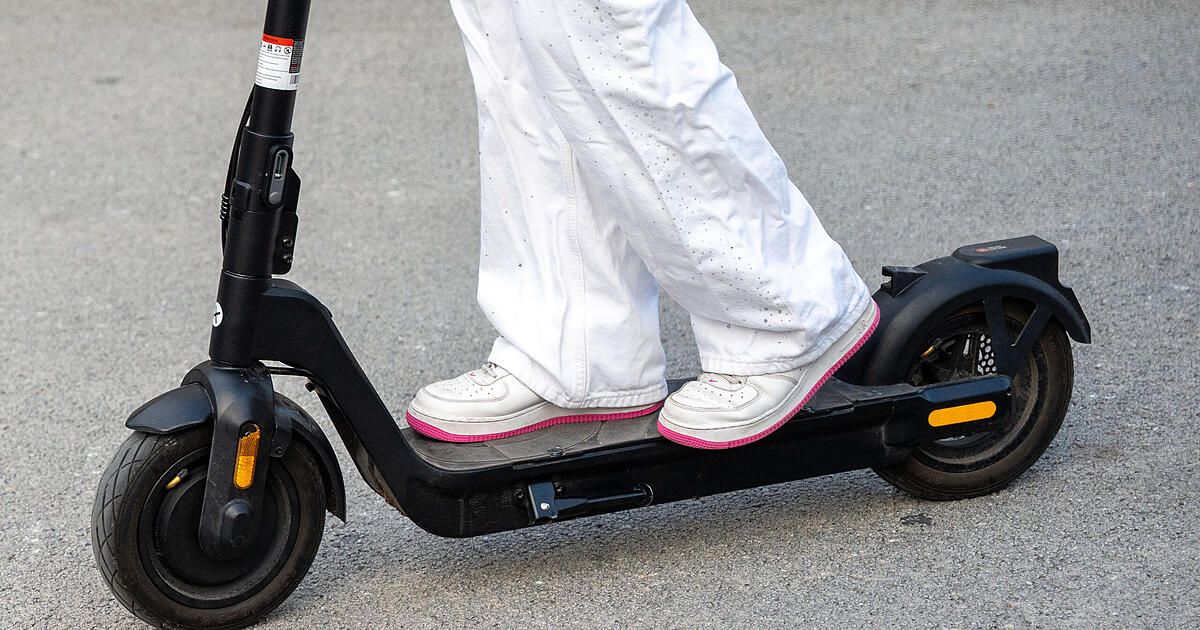 Eine Person steht auf einem schwarzen Elektroroller, trägt weiße Hosen und Schuhe mit rosa Sohlen. Der Roller hat einen schwarzen Lenker und ein gelbes Licht.