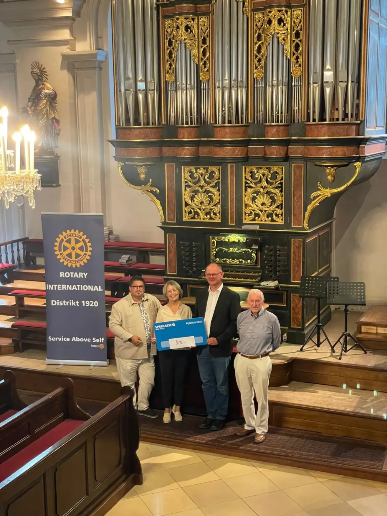 Vier Personen stehen vor einer Kirchenbühne und halten einen großen Scheck. Sie lächeln und scheinen für ein Foto zu posieren. Hinter ihnen steht eine große Orgel.