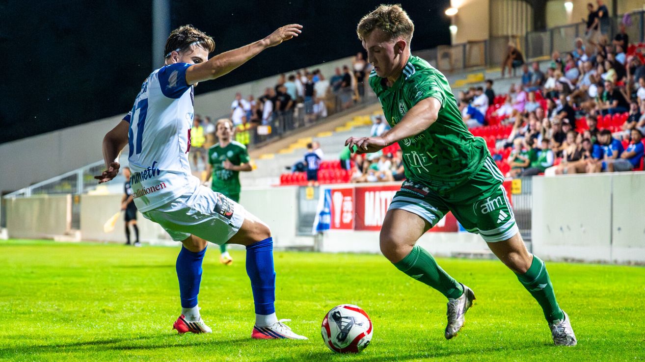 Two soccer players compete on a field; one in white and blue, the other in green. The player in green is dribbling the ball.
