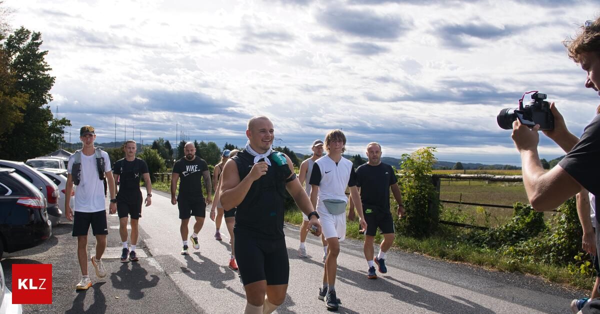 Eine Gruppe von Männern joggt auf einer Straße mit einem bewölkten Himmel darüber. Ein Mann lächelt und hält eine Flasche.