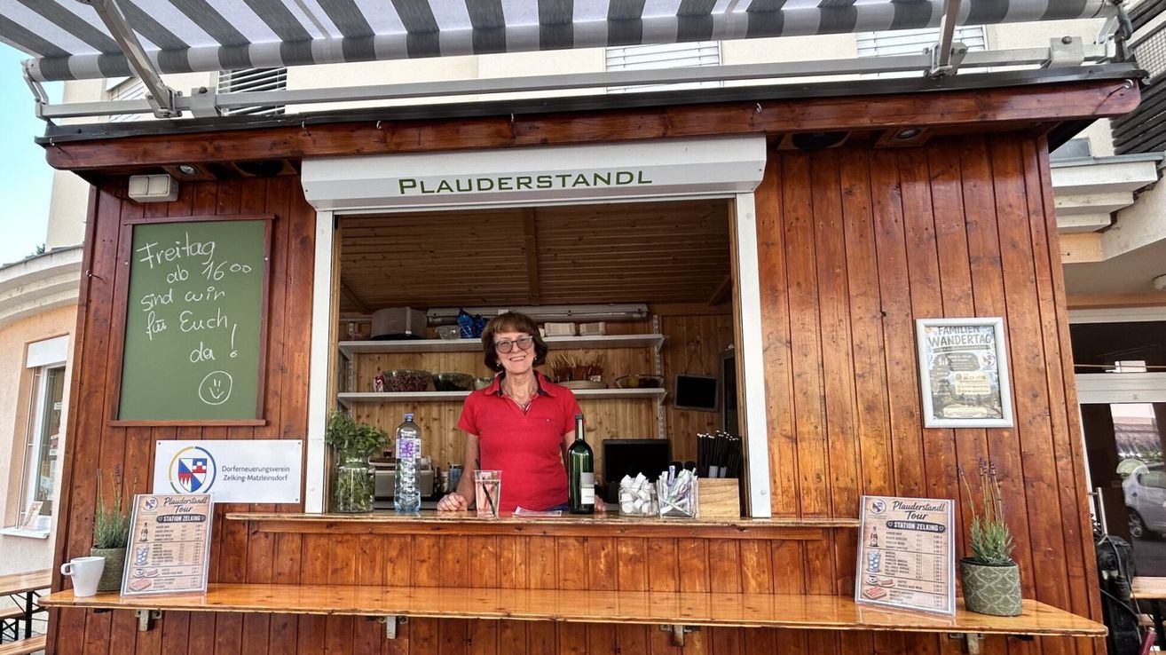 A woman stands behind a wooden counter in a small shop, wearing a red shirt and glasses. Bottles and items are on the counter, and a sign reads Plauderstandl.
