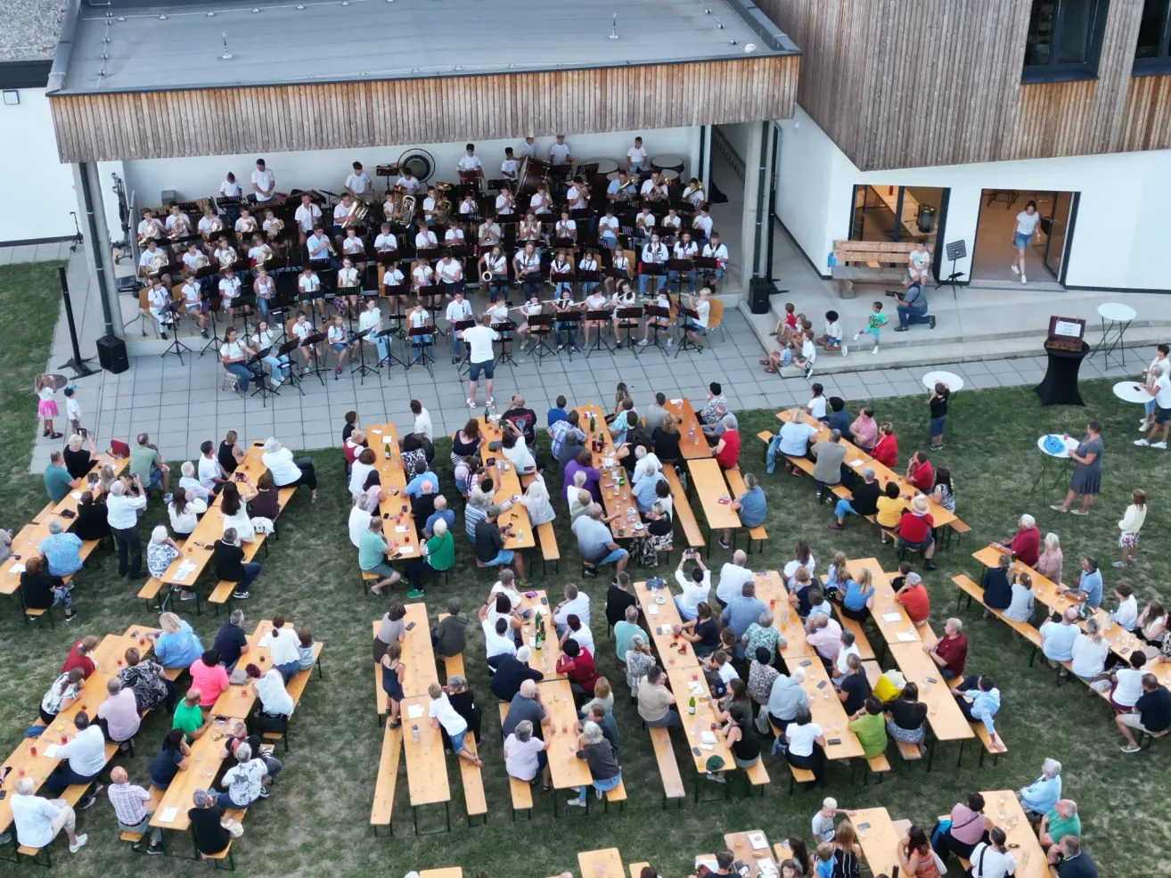 Eine große Gruppe von Menschen versammelt sich auf einem Freigelände, einige sitzen an Tischen, während andere stehen und eine Bandbesetzung beobachten. Die Band befindet sich auf einer Bühne mit Musikinstrumenten, und das Publikum sitzt in Reihen auf Bänken. Die Szene spielt sich auf einem Rasenfeld ab, mit einem Gebäude im Hintergrund.