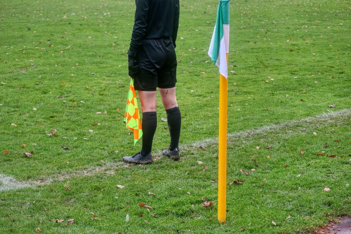 A soccer referee stands on a grassy field, holding a yellow and orange flag. The field has white markings and a yellow goalpost.