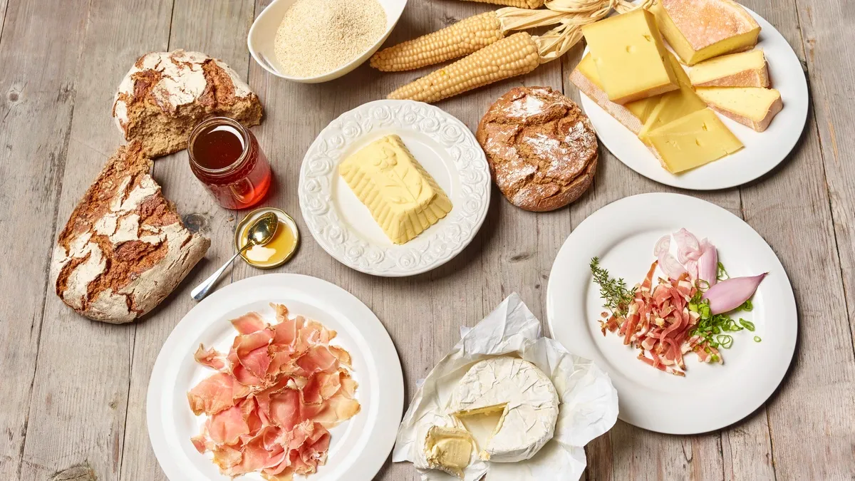 A wooden table is set with various foods including cheese, bread, honey, corn on the cob, and sliced ham.