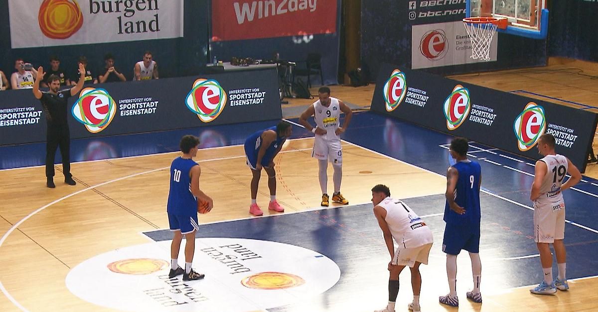A basketball game is in progress on an indoor court. Players in blue and white jerseys are standing and looking at the referee. The court has a logo on the floor and a basketball hoop. Advertisements are on the walls.