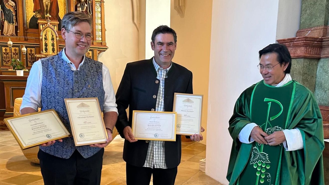 Drei Männer stehen in einer Kirche, lächeln und halten gerahmte Urkunden in den Händen. Der Mann in der Mitte trägt einen schwarzen Anzug, der Mann auf der linken Seite trägt eine Weste und eine Brille. Der Mann auf der rechten Seite trägt einen grünen Talar.