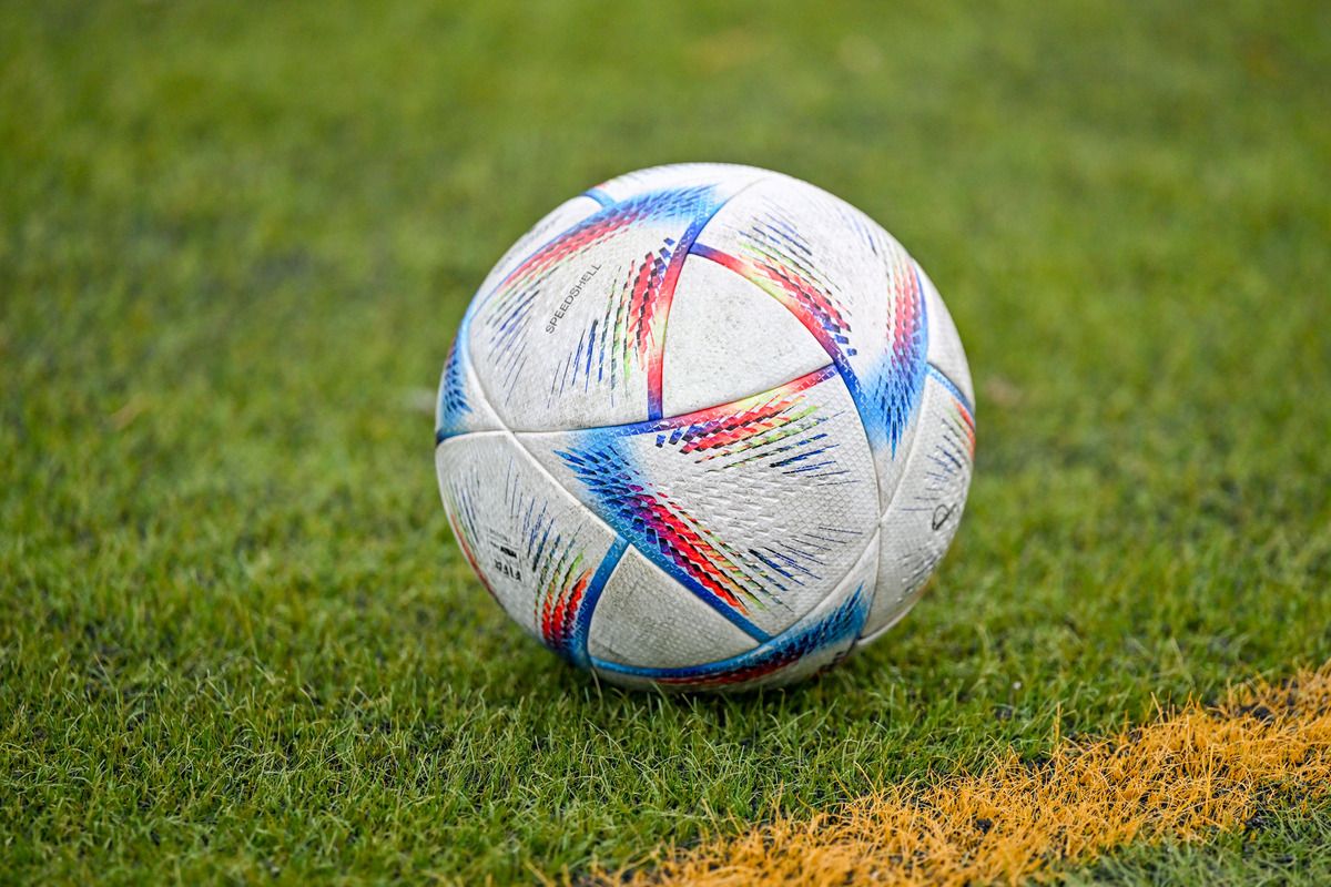 Ein Fußball mit weißem und mehrfarbigem Design liegt auf einem grünen Feld. Der Ball hat ein Markenlogo und eine gelbe Linie auf dem Gras.