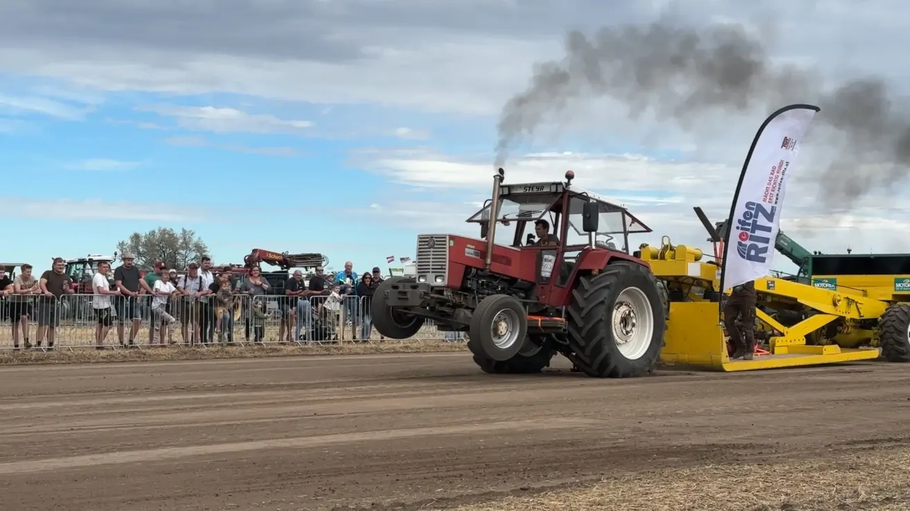 Ein roter Traktor fährt auf einer Erdstrecke und gibt Rauch ab. Ein Publikum beobachtet hinter einem Zaun.