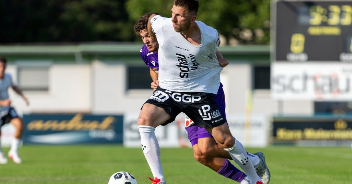 Zwei Fußballspieler konkurrieren auf einem Feld. Ein Spieler in Weiß jagt dem Ball nach, während der andere in Lila versucht, ihn zu blockieren. Der Spieler in Weiß trägt ein Shirt mit dem Wort 'Schrad' und Shorts mit 'GGP'. Der Hintergrund zeigt einen Zaun mit Werbung.