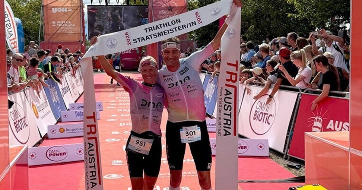 Zwei Athleten stehen auf einem roten Teppich und halten ein weißes Banner mit der Aufschrift 'TRIATHLON 2026 STATTSMEISTER/IN'. Sie tragen beide Radfahreranzüge und Stirnbänder. Hinter ihnen jubelt eine Menge Menschen. Im Hintergrund befindet sich eine große Leinwand und ein Banner mit dem Wort 'OMNI Biotic'.
