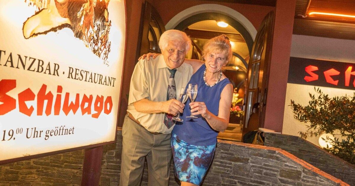 An elderly couple stands outside a restaurant, smiling and holding wine glasses. The sign behind them reads 'RESTAURANT WAGO' in red and white.