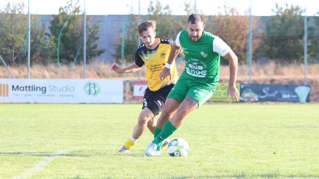 Zwei Fußballspieler sind auf einem Feld. Einer ist in Gelb und Schwarz, der andere in Grün und Weiß. Sie jagen einem Fußball nach.