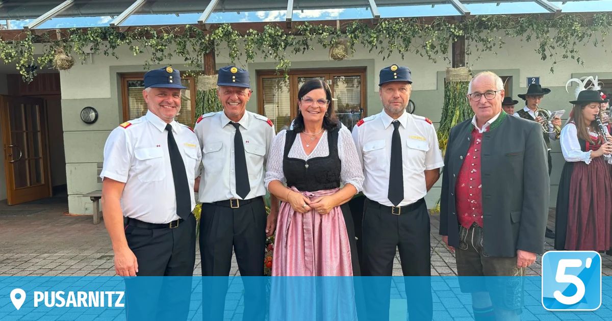 Fünf Personen in Uniformen und traditioneller Kleidung stehen vor einem Gebäude mit einer grünen Pflanze darüber.