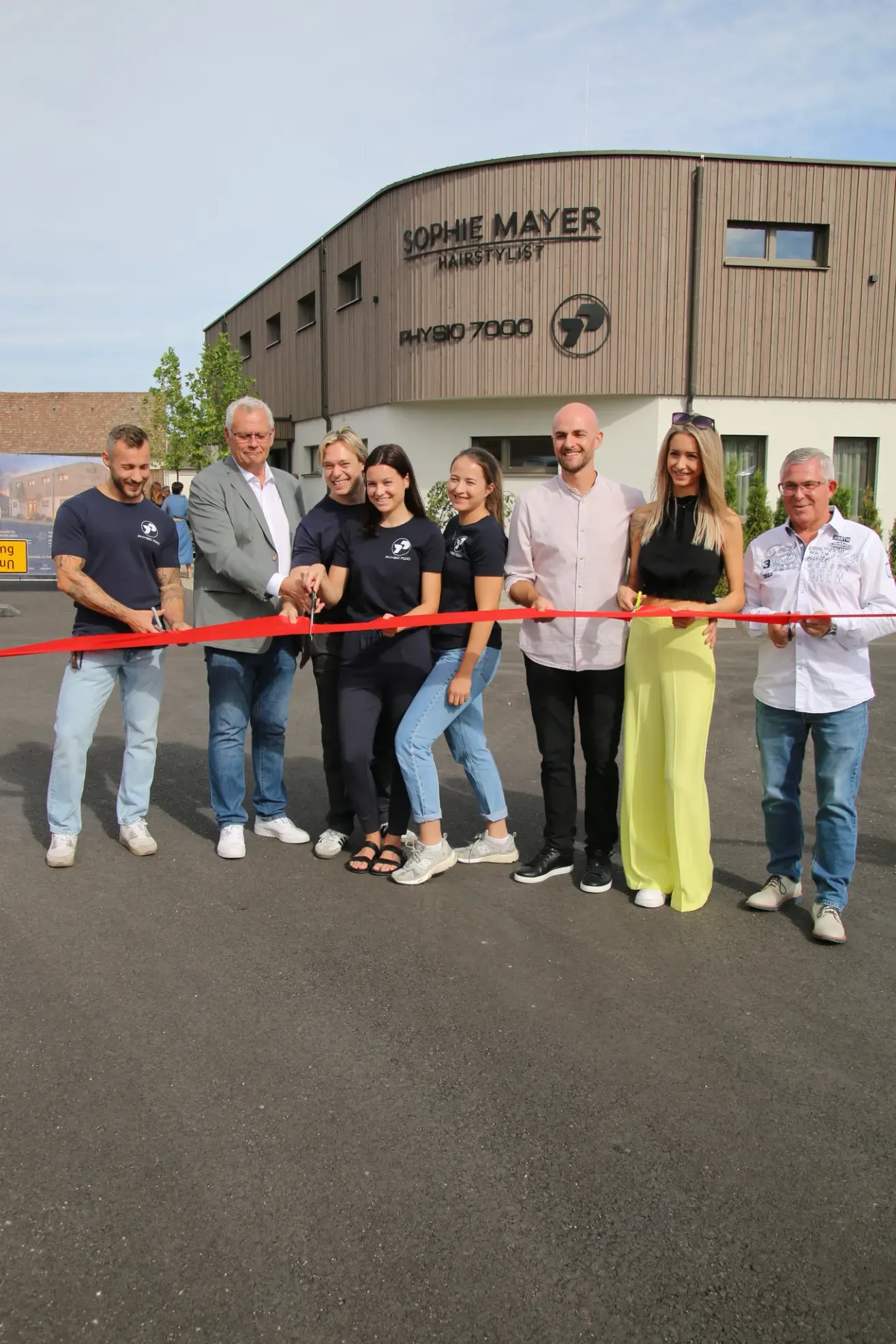 A group of people are gathered in front of a building, holding a red ribbon for a ribbon-cutting ceremony.