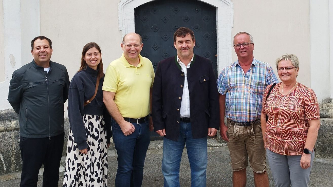 Four people stand in front of a building with a black door. The woman wears a black and white dress, the man on the left a yellow shirt, the man in the middle a black jacket, and the man on the right a plaid shirt.