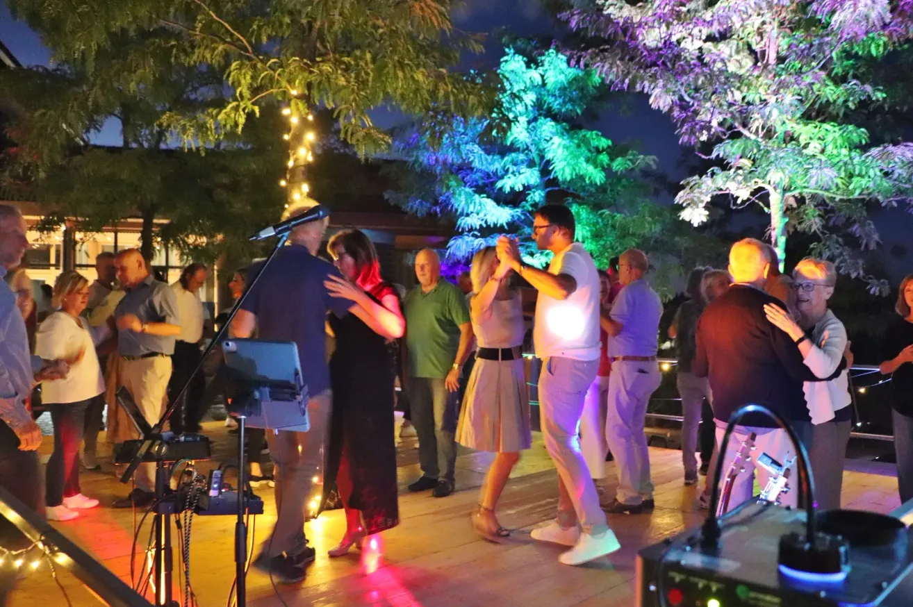 A group of people dance on a wooden deck at night, illuminated by colorful lights and trees. A man sings into a microphone.