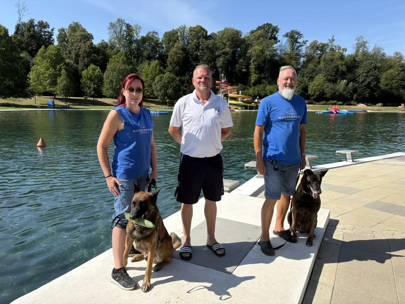 Three people and two dogs stand on a dock by a lake, smiling for the camera. The man in the middle wears a white polo, while the others wear blue shirts. The dogs are leashed and appear calm.