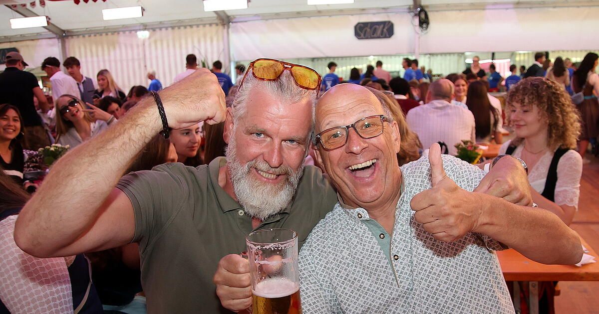 Two older men are posing for a photo at a crowded outdoor event, one giving a thumbs up and the other holding a glass of beer.