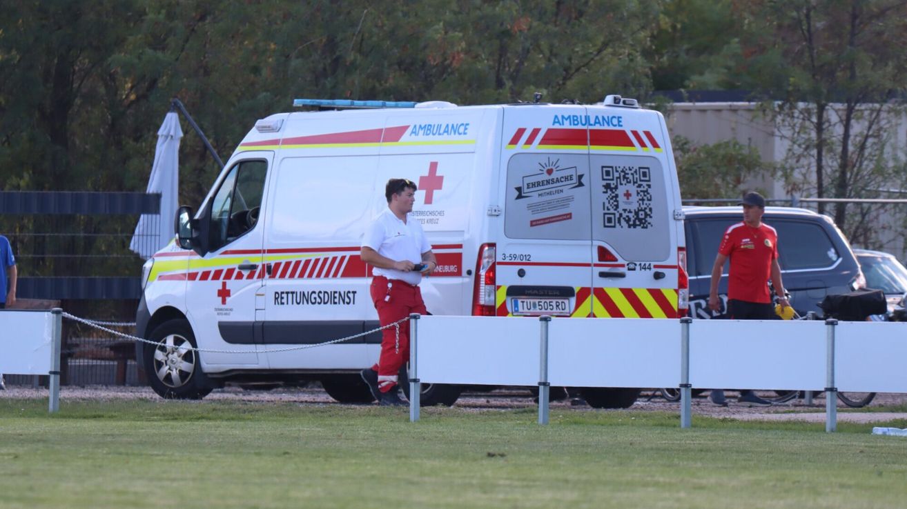Ein Mann in roter und weißer Uniform steht neben einem Krankenwagen mit rotem Kreuz und QR-Code auf der Rückseite.