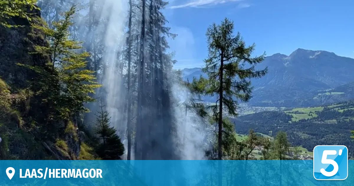 A mountain landscape with a waterfall cascading through a dense forest. The water is white and misty, and the trees are green. The sky is clear and blue.