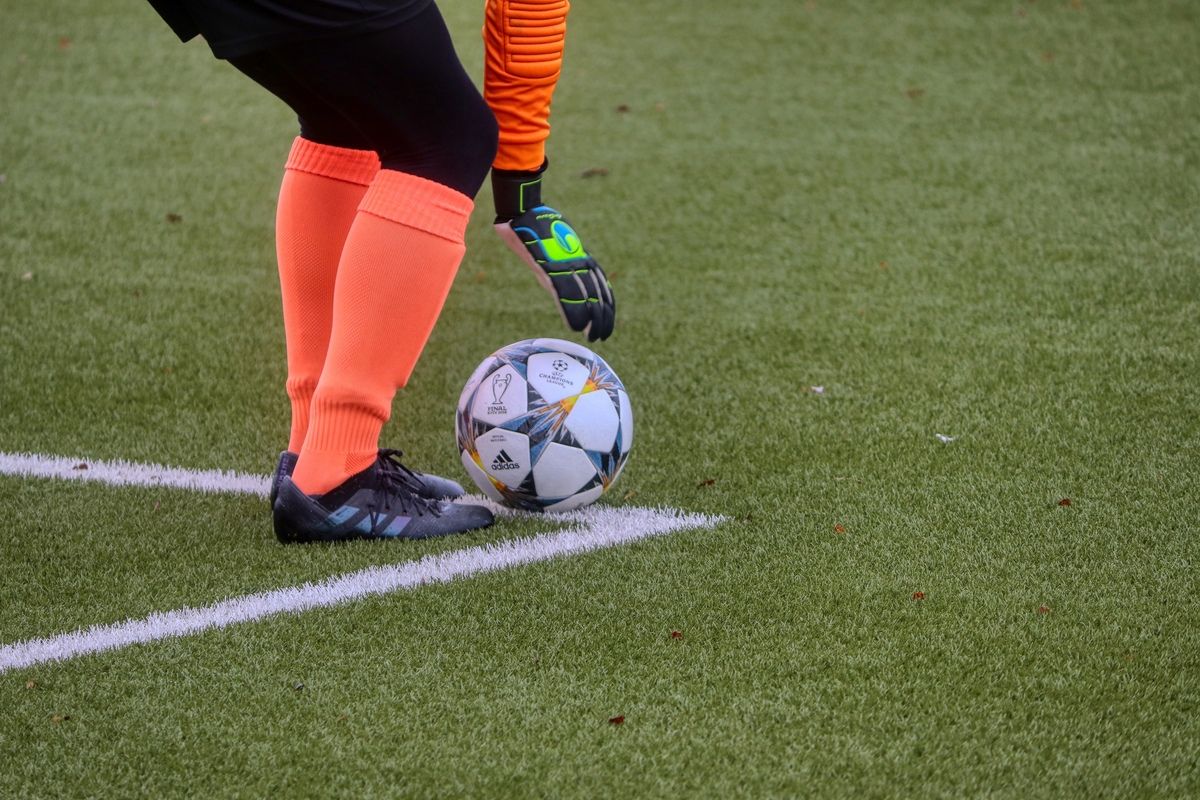Ein Fußballspieler in orangefarbenen Socken steht auf einem Feld und ist bereit, einen Ball zu schießen. Der Ball ist Adidas-markiert, und der Spieler trägt schwarze Schuhe.