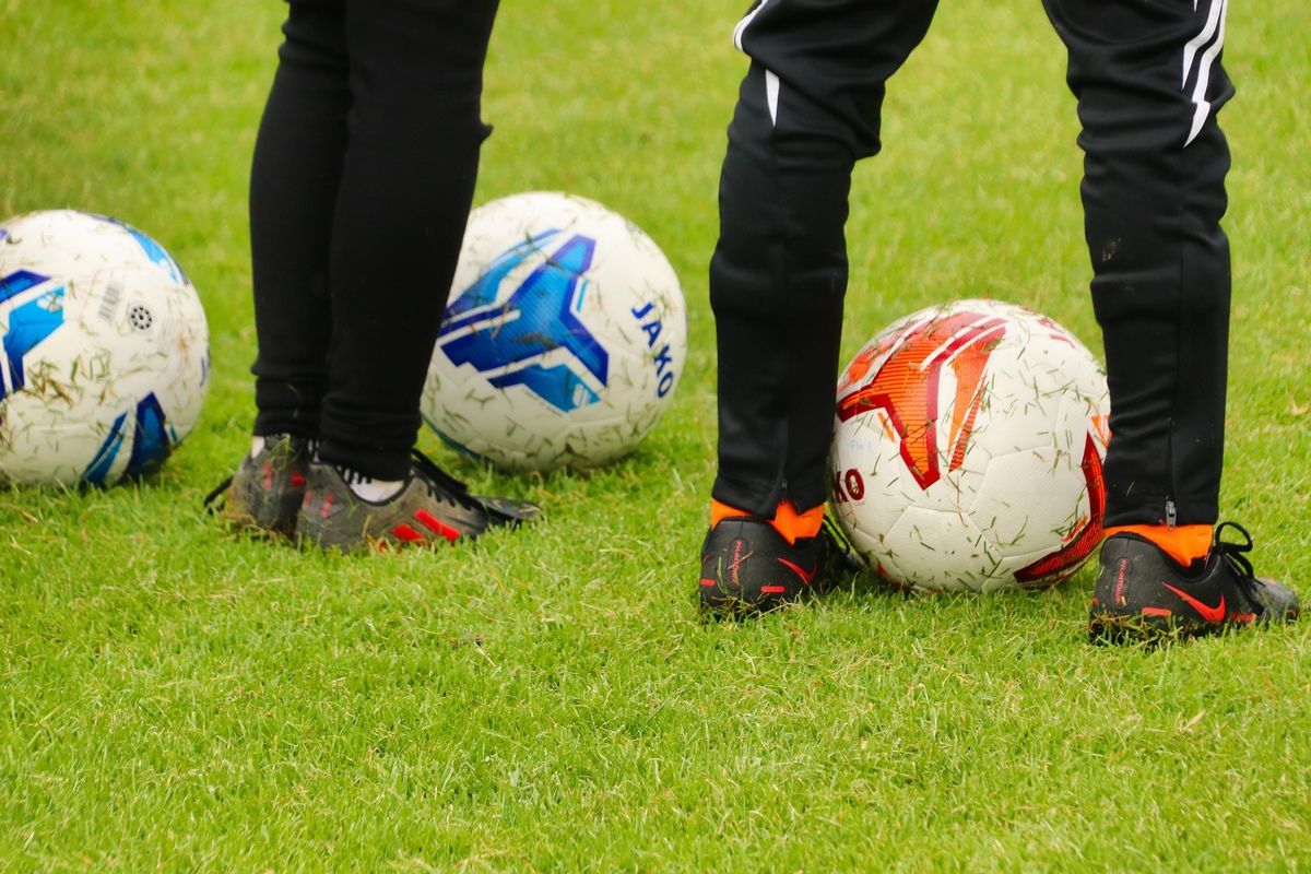 Two soccer players stand on a grassy field, each with a soccer ball in front of them. One ball has a blue and white design, the other red and white.