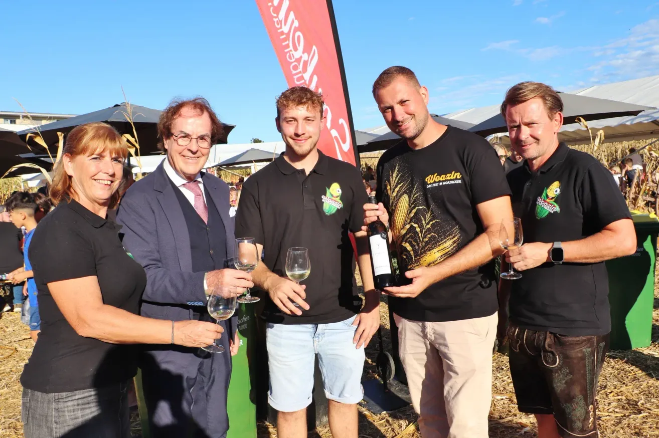 Four men stand together, smiling, holding wine glasses and a bottle. They appear to be at an outdoor event. Behind them is a banner with the word 'Weizen'.