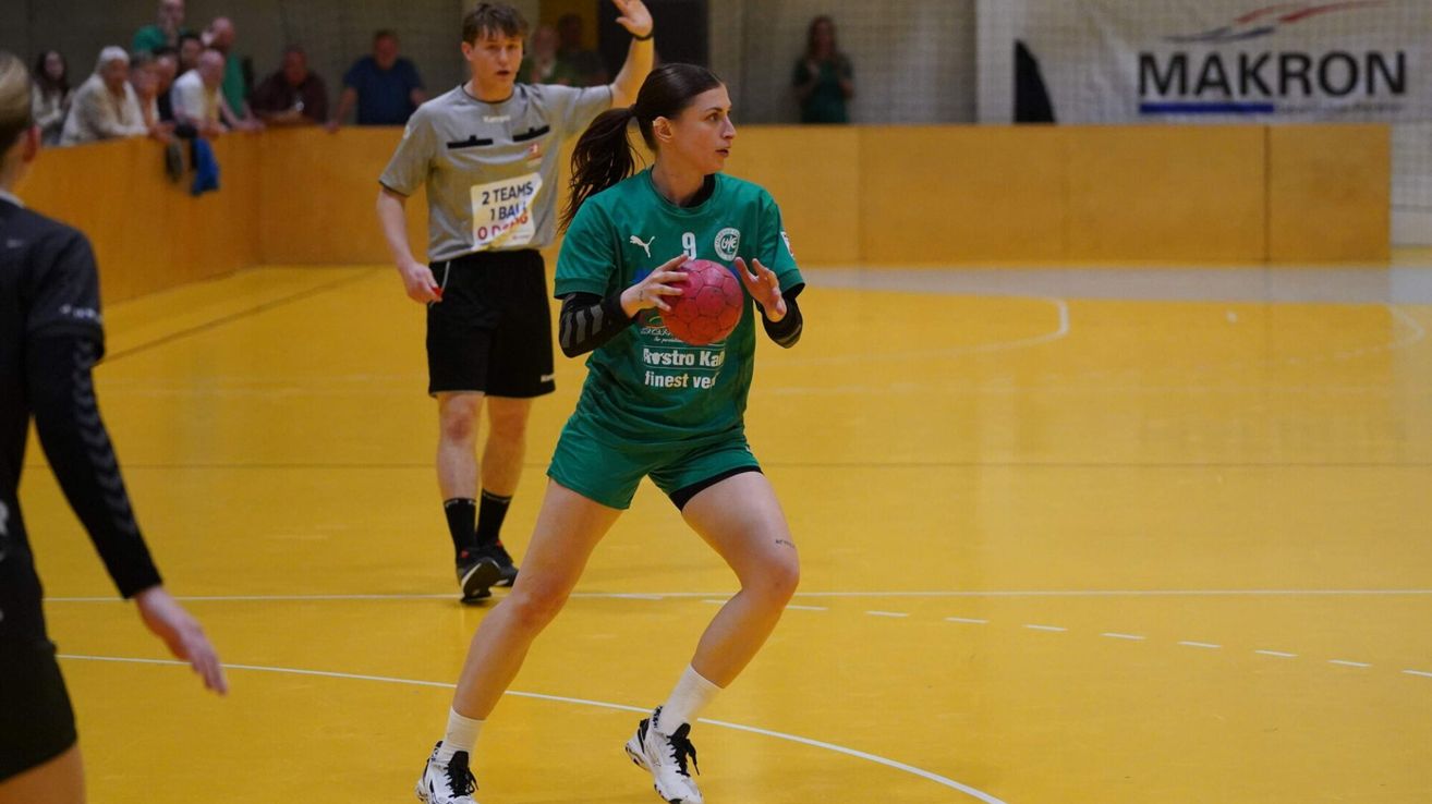 A female athlete in green uniform holds a red ball, playing handball on a yellow court with a referee behind her.