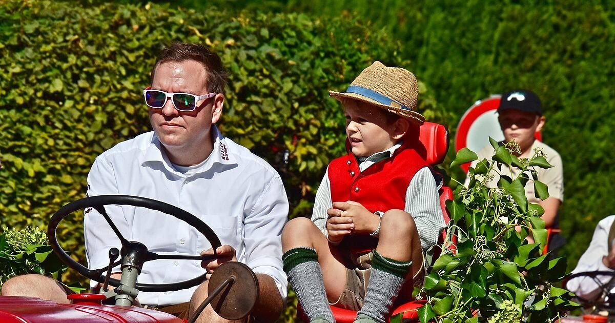A man and a young boy are sitting in a tractor, with the boy wearing a hat and red vest. The man is wearing sunglasses and holding the steering wheel. They are surrounded by green plants.