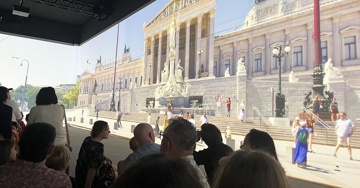 Eine große Gruppe von Menschen schaut auf eine große Leinwand, die eine Ansicht eines historischen Gebäudes zeigt.