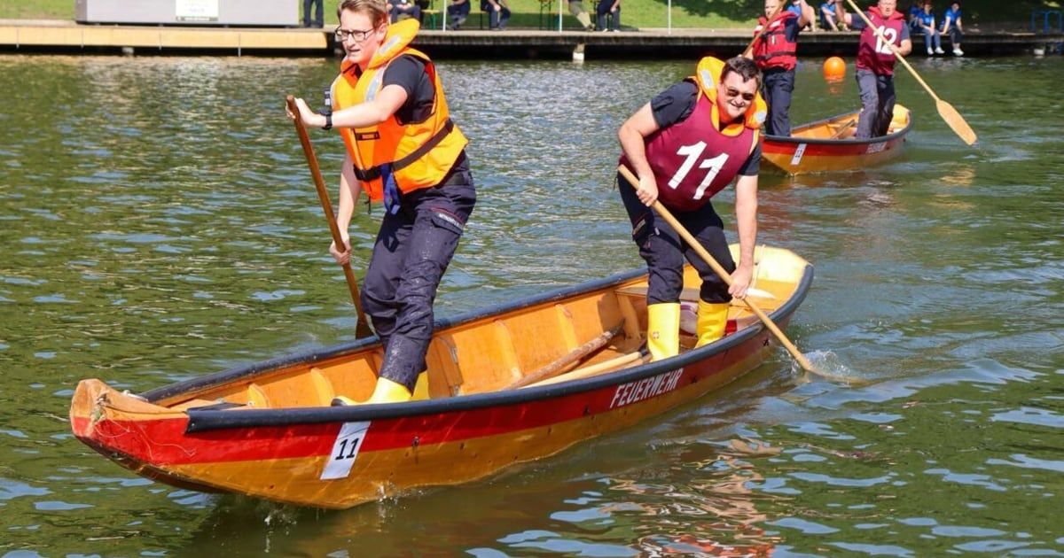 Two people in life jackets paddle a boat labeled 'Feuerwehr' on a lake with other boats in the background.