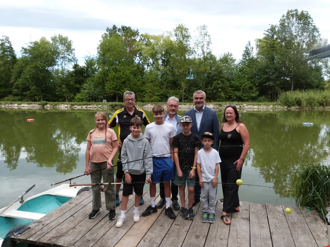 A group of people, including adults and children, stand on a wooden dock by a lake, holding fishing rods. They are smiling and appear to be posing for a photo.