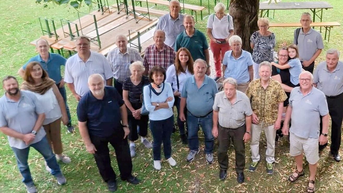 A group of people, mostly elderly, are standing in a park with benches and a tree in the background. They are dressed casually and appear to be posing for a photo.