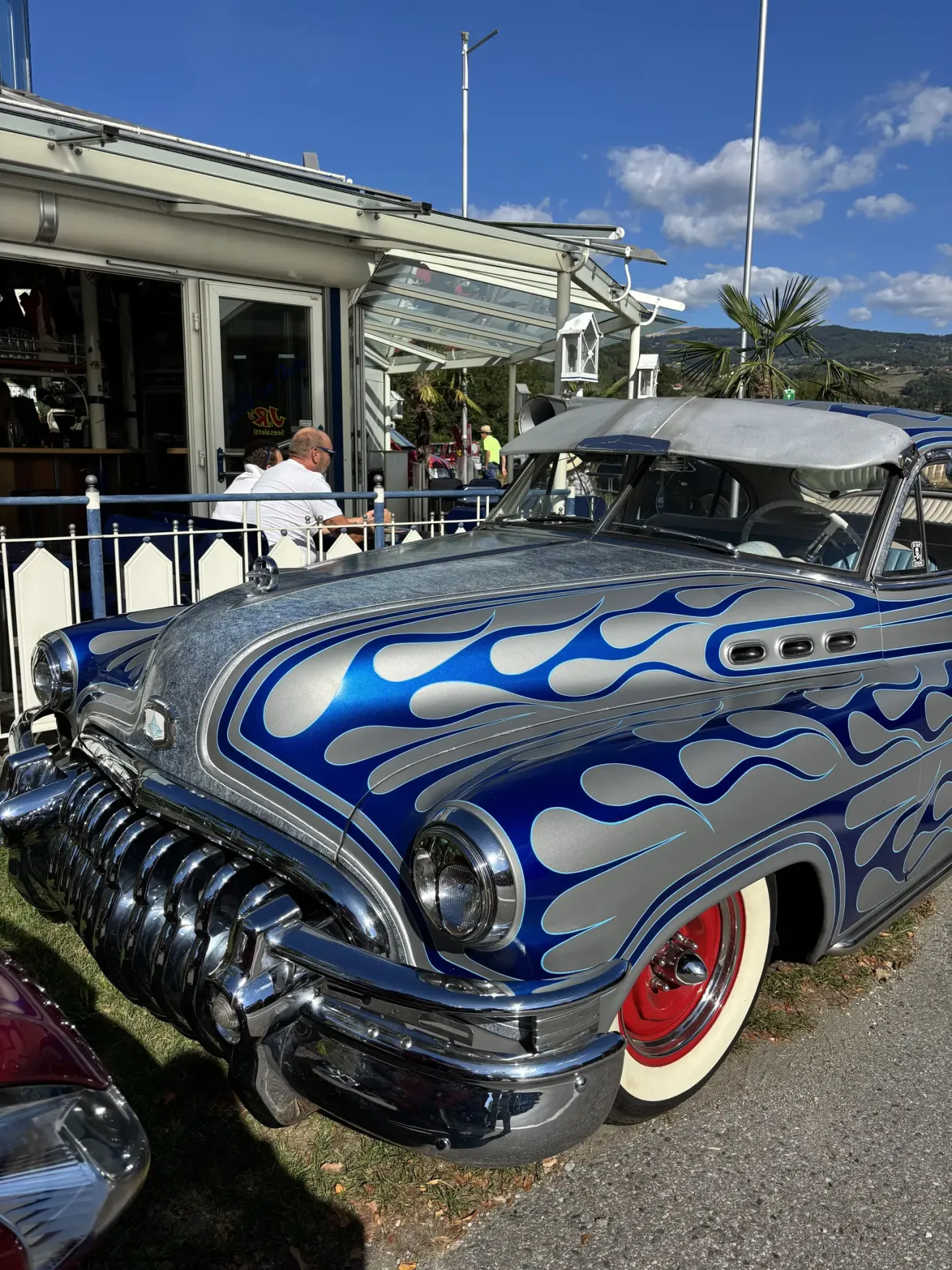 A vintage blue and silver car with flame-like designs is parked in front of a restaurant. A man is sitting on a bench outside.