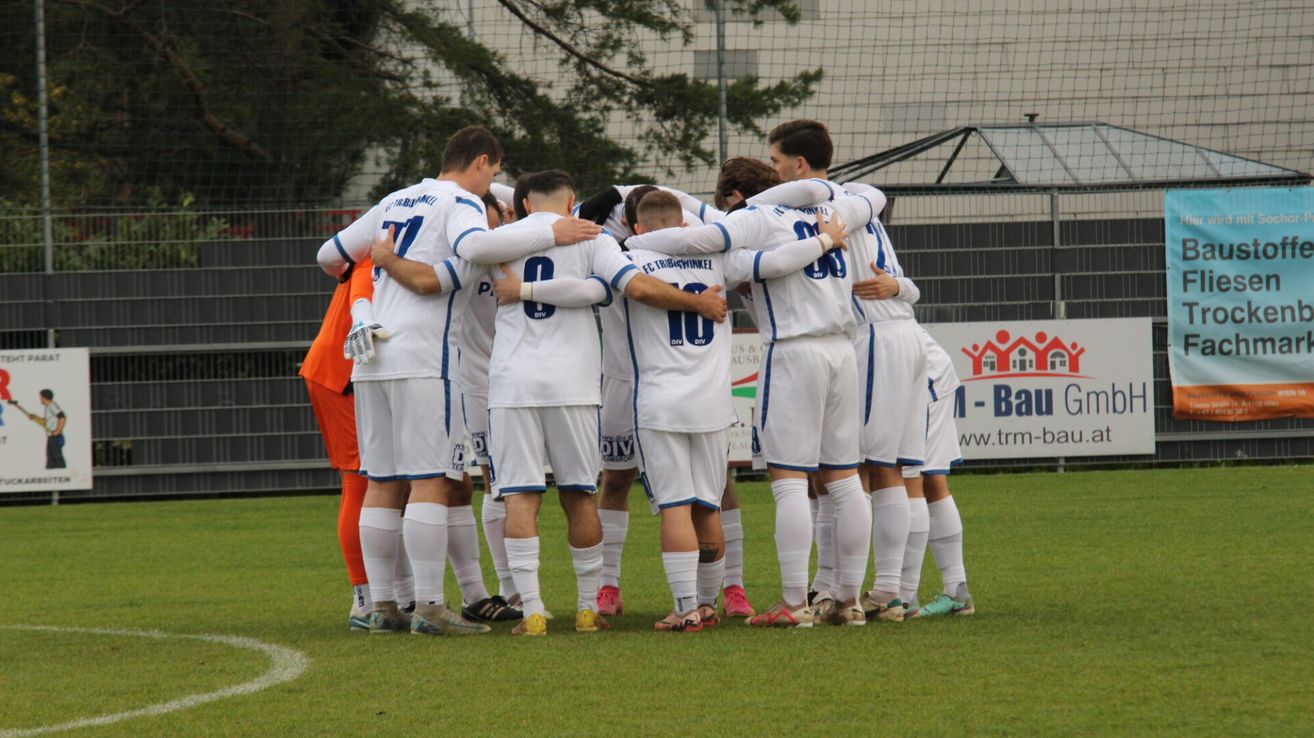 Eine Gruppe von Fußballspielern in weißen Trikots steht auf einem Feld versammelt und bespricht wahrscheinlich ihre Strategie. Der Spieler mit der Nummer 7 auf seinem Trikot gibt einen Daumen hoch.