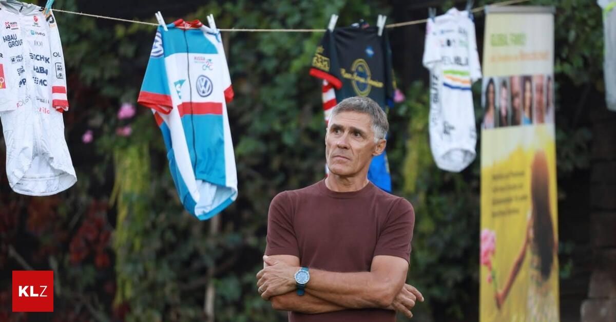 An older man stands with folded arms in front of a clothesline with various cycling jerseys hanging.