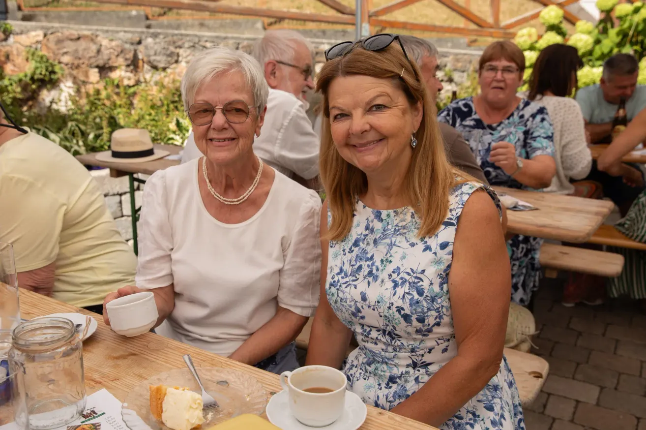 Two women sitting at a table, smiling and holding cups. One woman wears a pearl necklace and the other has a floral dress. Behind them, other people are seated at tables.
