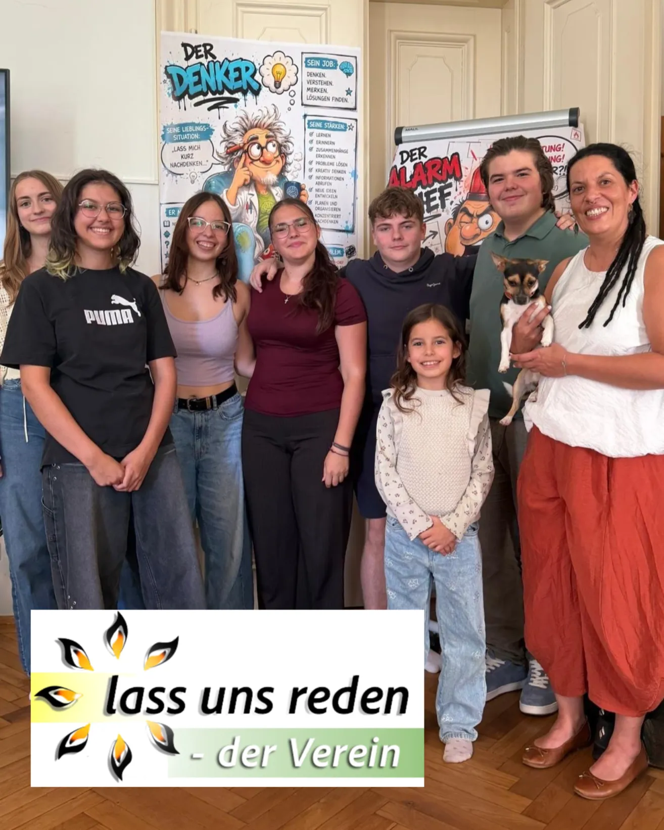 A group of people, including adults and children, are posing for a photo in a room with a poster in the background. The woman on the right is holding a small dog.