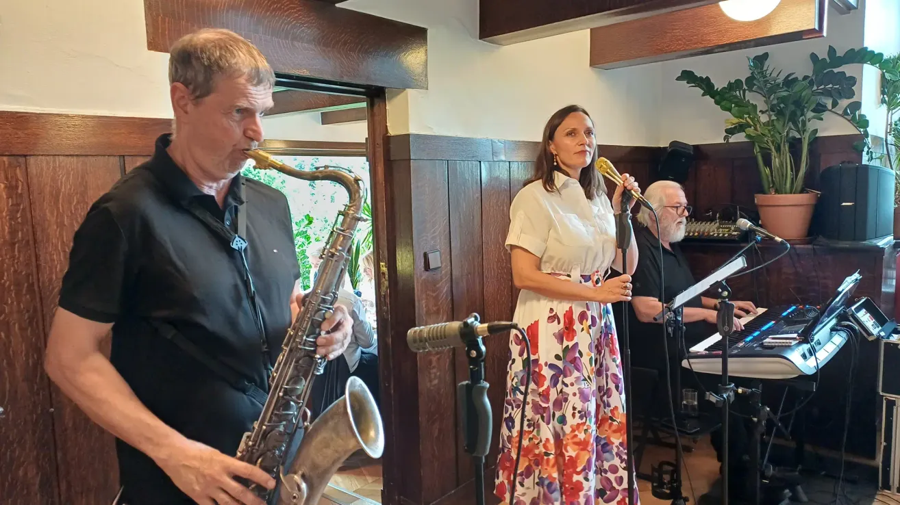 A man plays the saxophone while a woman sings into a microphone on stage. Behind them, another man plays the piano. The setting appears to be a small, intimate performance venue.