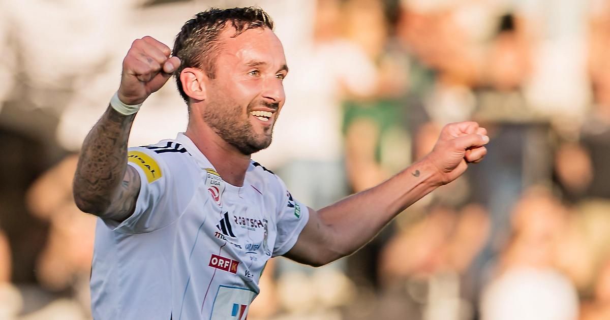 Ein Fußballer feiert mit erhobenen Armen, trägt ein weißes Trikot mit verschiedenen Logos und ein Tattoo auf seinem rechten Arm.