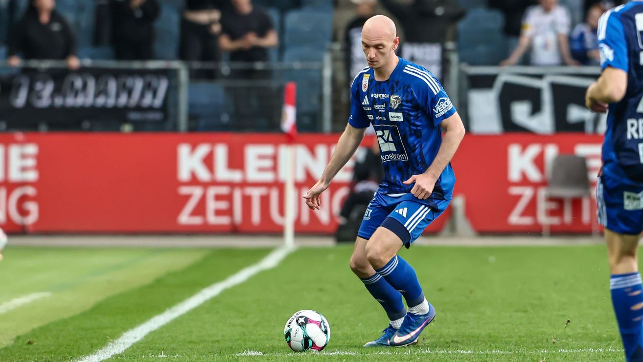 Ein Fußballspieler in blauer Uniform dribbelt einen Fußball auf einem Feld mit einem roten Banner im Hintergrund.